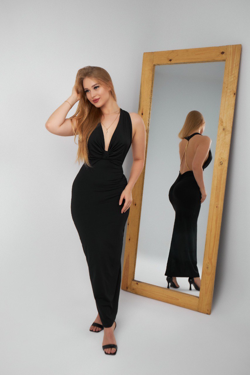 Woman in a black dress standing next to a full-length mirror on a gray background